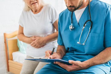 Wall Mural - cropped shot of doctor writing on clipboard while sick senior woman sitting on hospital bed