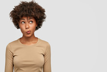 Young Lovely Female Has Dark Skin, Clueless And Unaware Expression, Purses Lips As Being Questionned, Looks With Puzzlement Upwards, Isolated Over White Background With Copy Space On Right Side