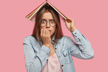 Wall Mural - Anxious nervous Caucasian female bites finger nails, holds book above head, worries before passing exam, poses against pink backgroud. Student looks nervously at camera. People and education concept