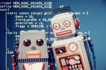two tin toy robots standing still like in a selfie shot. background: computer source code (public do