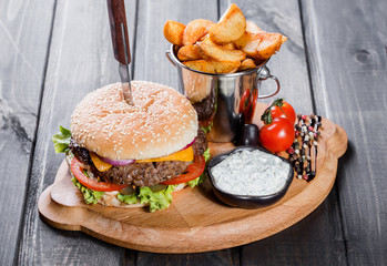 Canvas Print - Hamburger with fresh vegetables, cheese, sauce and fries on cutting board on dark wooden background. Burger with knife