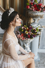 Beautiful and fashionable brunette model girl with bright makeup and diadem on her head, in stylish lingerie and in a peignoir sits on the bed and posing in luxury vintage interior