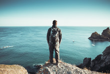 A Rear View Of A Male Backpacker Or Hiker Standing On A High Clifftop And Overlooking A Vast Ocean At Kynance Cove In Cornwall, UK