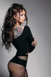 Back view of sexy and beautiful girl with seductive figure and colorful tattoo on shoulder, posing at studio on grey background. Sensuality model wearing black casual blouse with closed eyes.