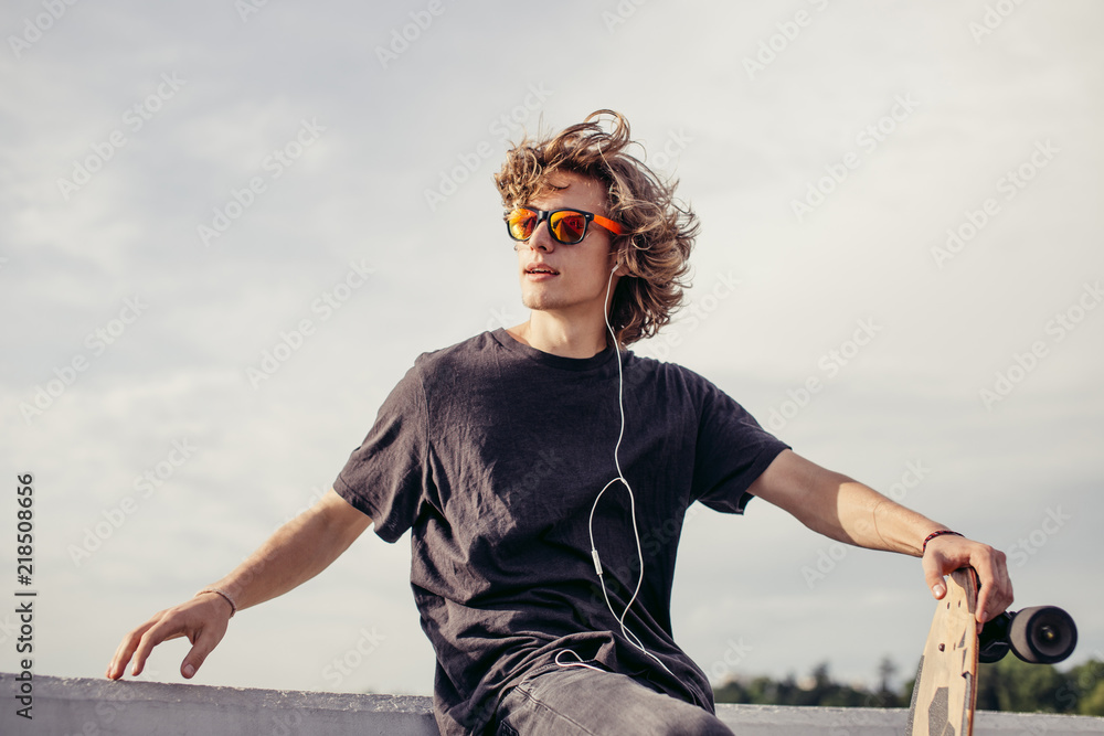 stylish young man with skateboard listening music with headphones , near concrete wall - obrazy, fototapety, plakaty 