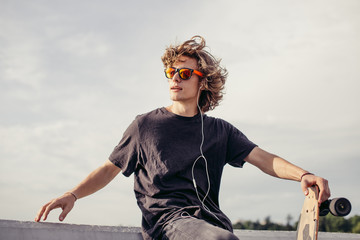 stylish young man with skateboard listening music with headphones , near concrete wall