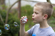 © bilanol - Profile of cute handsome small blond child boy with funny serious expression blowing colorful transparent soap bubbles outdoors on blurred green summer background. Joy of careless childhood concept.