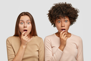 Wall Mural - Horizontal shot of beautiful scared two multiethnic women with stunned expressions, keep hands near widely opened mouthes, react on fake news, stand next to each other, isolated over white background.