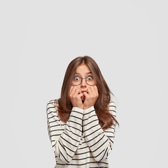 Poster - Frightened beautiful young woman bites finger nails, looks anxiously at camera, dressed in fashionable clothes, has appealing look, stands against white background. People, emotions concept.
