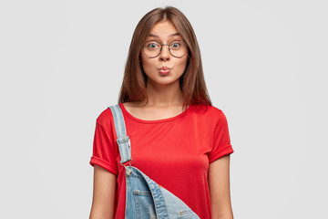 Poster - Lovely cute young female with friendly expression, makes grimace, pouts lips, dressed in stylish red t shirt and overalls, poses against white background. Adorable student ready for walk outside