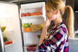 © nenetus - Pretty young woman hesitant to eat in front of the fridge in the kitchen.