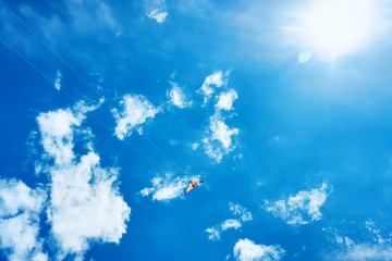 A large kite floats high in the sky among the clouds against the blue sky and the bright summer sun and it is controlled by two white ropes going to the ground