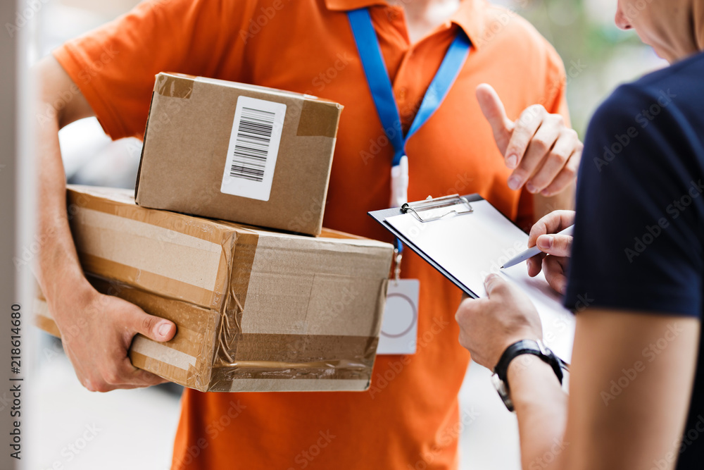 A person wearing an orange T-shirt and a name tag is delivering a parcel to a client, who is putting his signature on the receipt. Friendly worker, high quality delivery service. - obrazy, fototapety, plakaty 