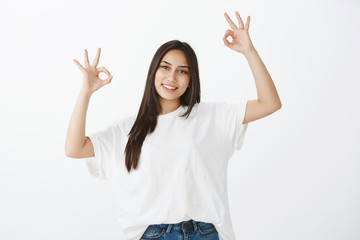 No worried, everything ok. Portrait of positive grinning female student in white t-shirt, raising hands in okay or great gesture, smiling happilly while liking idea or giving approval over gray wall