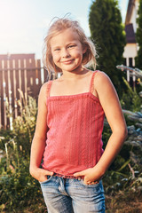 Portrait of a smiling cute girl with blonde hair holds hands on pockets while standing in the backyard.