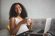© Anatoliy Karlyuk - People, leisure, technology and gadgets concept. Portrait of stylish beautiful young African American woman with wavy hair enjoying slow morning, having cup of coffee and surfing internet using laptop
