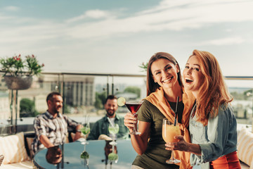 Happy emotions. Laughing young ladies posing with alcohol drinks at rooftop cafe while gentlemen sitting at the table