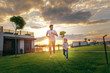 Full length portrait of beaming unshaven dad running with laughing child on green grass during sunny evening