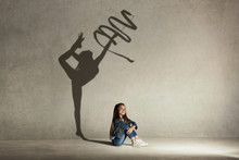 Baby Girl Dreaming About Gymnast Profession. Childhood And Dream Concept. Conceptual Image With Shadow Of Female Gymnast On The Studio Wall