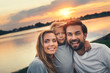 © Yakobchuk Olena - Portrait of outgoing wife and glad bearded husband hugging smiling kid opposite beautiful sunset under river in outside. They laughing and looking at camera