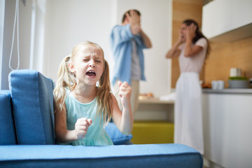 Wall Mural - Little capricious girl expressing her displeasure by loud voice and hitting armchair with shocked parents on background