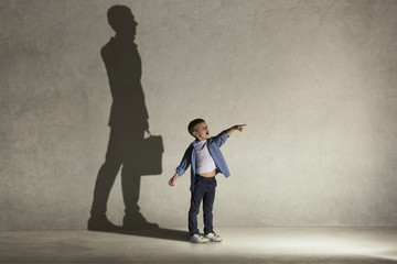 The little boy dreaming about businessman or diplomat profession. Childhood and dream concept. Conceptual image with boy and shadow of man in suit on the studio wall