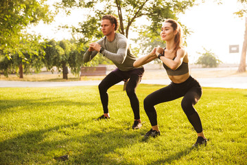 Wall Mural - Image of young caucasian sporty man and woman 20s in tracksuits, doing workout and squatting together in green park during sunny summer day