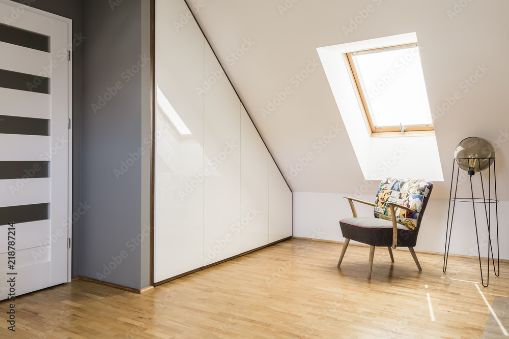 Lamp next to armchair on wooden floor in white attic interior with door and window. Real photo - obrazy, fototapety, plakaty 