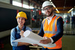 © Seventyfour - Portrait of modern bearded engineer wearing hardhat holding blueprints smiling cheerfully while discussing production with female worker at factory, copy space
