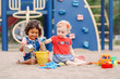 © anoushkatoronto - Sandy ground. Two cute Caucasian and hispanic latin babies children sitting in sandbox playing with plastic colorful toys. Little girls friends having fun together on playground.