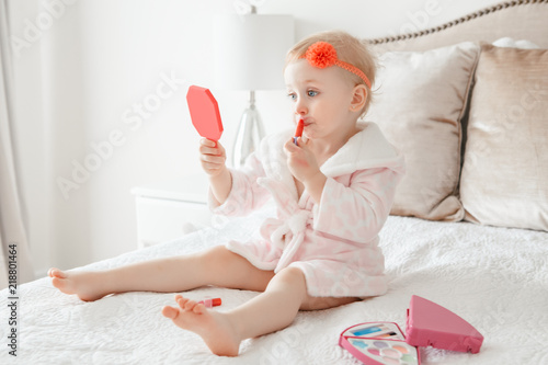 Cute Asorable Funny Caucasian Blonde Baby Girl 2 Years Old In Bathrobe Doing Applying Makeup Looking Into Mirror And Sitting On Her Bed Bedroom At Home Happy Childhood Concept Cute Asorable Funny Caucasian Blonde
