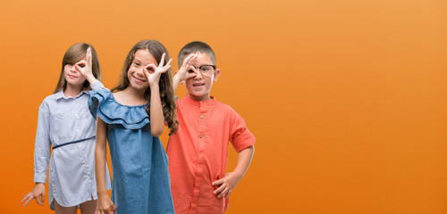 Sticker - Group of boy and girls kids over orange background with happy face smiling doing ok sign with hand on eye looking through fingers