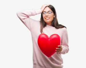 Poster - Young hispanic woman holding heart in love stressed with hand on head, shocked with shame and surprise face, angry and frustrated. Fear and upset for mistake.