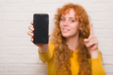 Sticker - Young redhead woman sitting over brick wall showing smartphone surprised with an idea or question pointing finger with happy face, number one