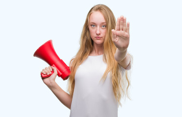 Sticker - Blonde teenager woman holding megaphone with open hand doing stop sign with serious and confident expression, defense gesture