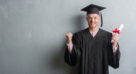 Young redhead man over grey grunge wall wearing graduate uniform holding degree screaming proud and celebrating victory and success very excited, cheering emotion