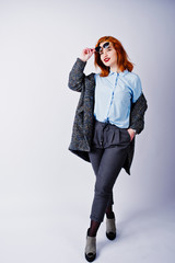 Portrait of a fantastic redheaded girl in blue shirt, grey overcoat posing with sunglasses in the studio.