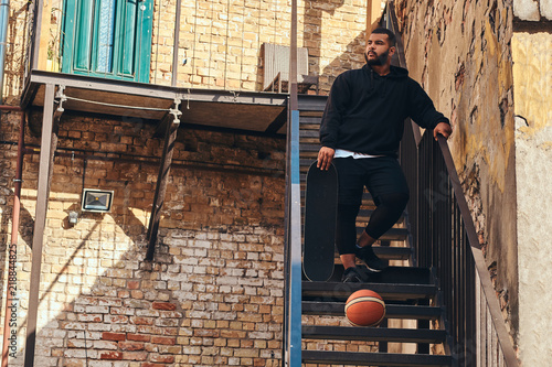 Photo  Dark skinned bearded hipster guy dressed in a black hoodie and sports shorts holds a skateboard while standing on stairs in ghetto