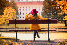 Attractive Young Woman With Red Hat Sitting On A Bench In Park And Enjoying A View On Castle At Autumn Season. Fashion Concept