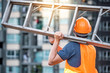 © zephyr_p - Young Asian maintenance worker man with orange safety helmet and vest carrying aluminium step ladder at construction site. Civil engineering, Architecture builder and building service concepts