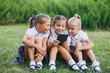 three little girls are sitting on the grass and reading an e-book. Pupils of primary school. Beginning of lessons. First day of fall.
