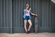 © Leika production - A pretty blond girl wearing checkered shirt and denim shorts is standing in front of the black wall with a black longboard next to her. Cool style.