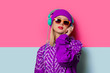 Young blonde girl in 90s sports jacket and hat on pink and blue background.