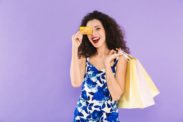 Poster - Portrait of beautiful woman 20s wearing dress holding credit card and paper shopping bags, standing isolated over violet background