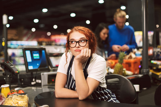 Young boring cashier in eyeglasses and striped apron dreamily looking aside while working in modern supermarket with customers on background
