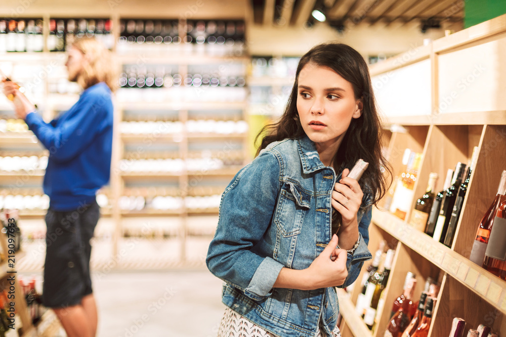 Young frightened woman in denim jacket trying steal bottle of wine in modern supermarket - obrazy, fototapety, plakaty 