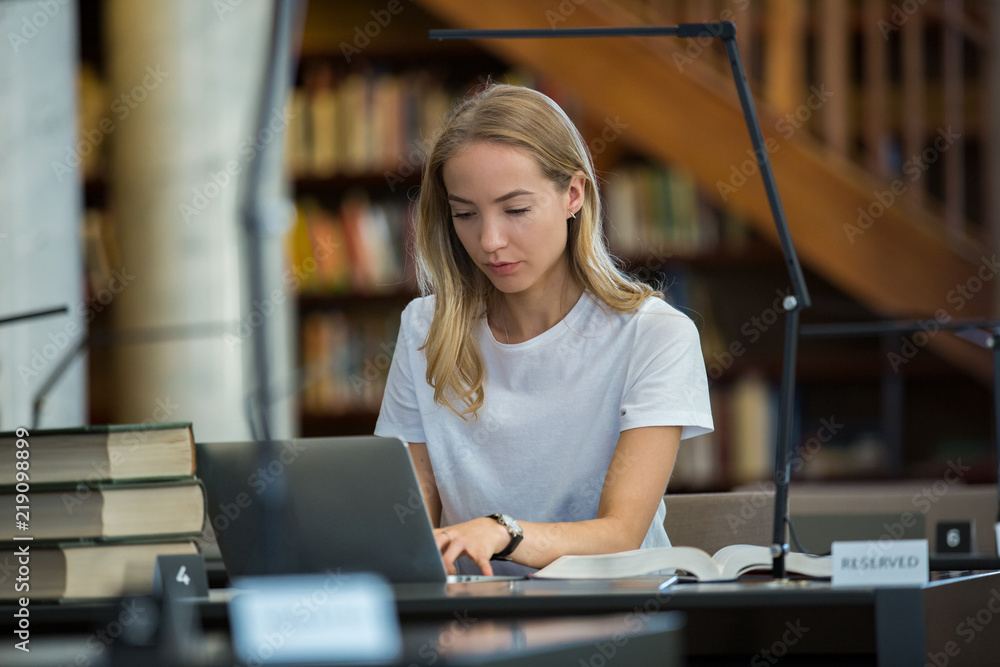 Young girl sitting at a desk in library, working at computer. Reading intently from screen, typing, studying and thinking. Higher education.  - obrazy, fototapety, plakaty 