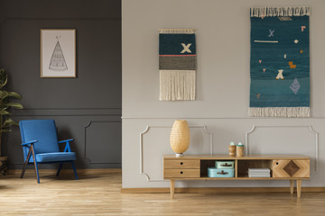 Lamp on wooden cupboard in living room interior with blue armchair under poster. Real photo