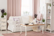 Real photo of bright provencal sitting room interior with white sofa, wooden coffee table on dirty pink carpet, rack with decor and fresh lavender