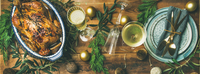 Christmas or New Year celebration table setting. Flat-lay of roast chicken or turkey, plates, silverware, glass and toy holiday decoration over rustic wooden background, top view, copy space